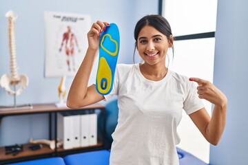Young hispanic woman holding shoe insole at physiotherapy clinic smiling happy pointing with hand and finger