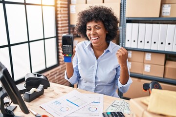 Black woman with curly hair working at small business ecommerce holding credit card and dataphone screaming proud, celebrating victory and success very excited with raised arm