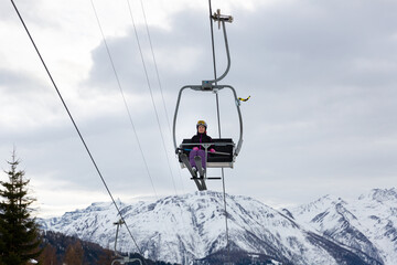 Fototapeta na wymiar Smiling woman skier in full ski equipment riding on chairlift, lifting to top of mountain to pistes on background of snowy peaks on cloudy winter day..