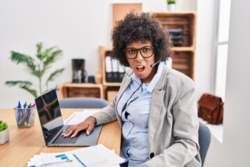 Black woman with curly hair wearing call center agent headset at the office in shock face, looking...