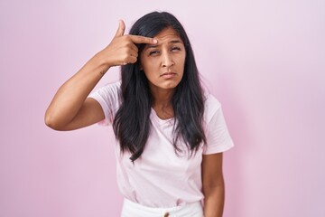 Young hispanic woman standing over pink background pointing unhappy to pimple on forehead, ugly...