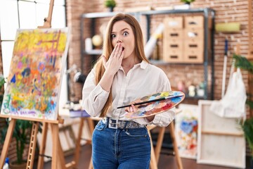 Young caucasian woman at art studio holding palette covering mouth with hand, shocked and afraid...