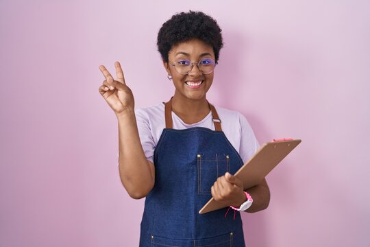 Young african american woman wearing professional waitress apron holding clipboard smiling with happy face winking at the camera doing victory sign with fingers. number two.