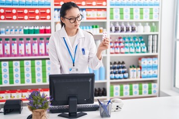 Young chinese woman pharmacist using computer holding pills tablet at pharmacy