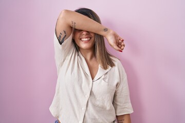 Blonde woman standing over pink background covering eyes with arm smiling cheerful and funny. blind concept.