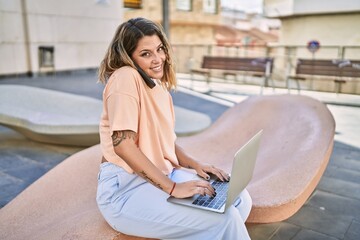 Young hispanic woman talking on the smartphone using laptop at street