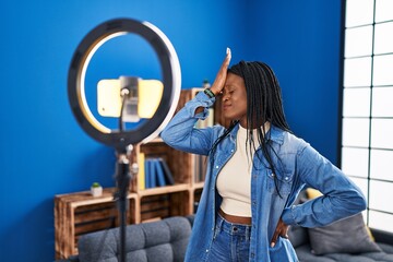African woman with braids recording vlog tutorial with smartphone at home surprised with hand on...