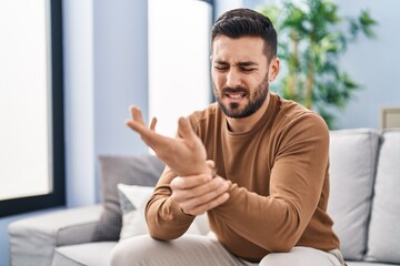 Young hispanic man suffering for wrist injury sitting on sofa at home
