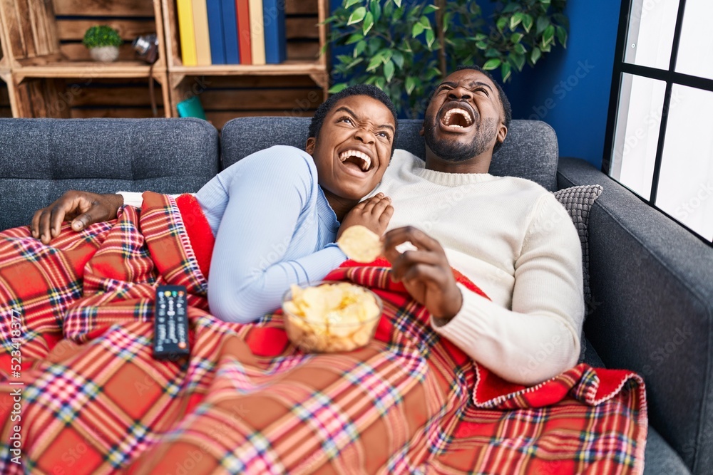 Wall mural Young african american couple sitting on the sofa watching tv angry and mad screaming frustrated and furious, shouting with anger looking up.