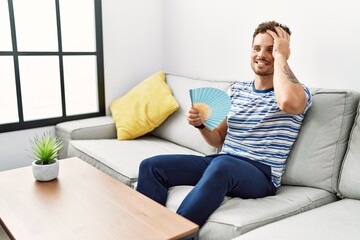 Young hispanic man smiling confident using hand fan at home