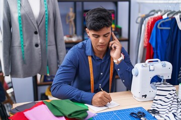 Young hispanic man tailor talking on smartphone writing on notebook at clothing factory