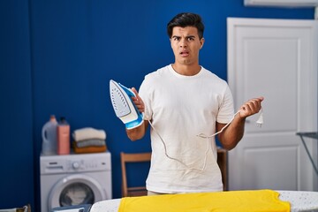 Hispanic man holding electric iron clueless and confused expression. doubt concept.