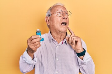 Senior man with grey hair holding medical asthma inhaler serious face thinking about question with...