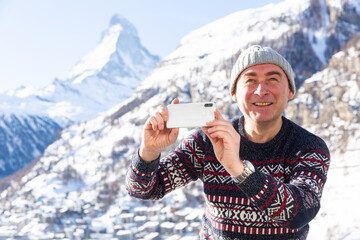 Cheerful excited traveler filming his hike in Swiss Alps on sunny winter day, capturing scenic mountain views from mobile phone