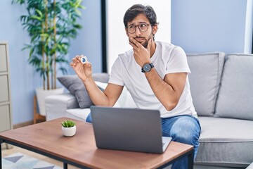 Handsome latin man holding virtual currency bitcoin using laptop serious face thinking about question with hand on chin, thoughtful about confusing idea