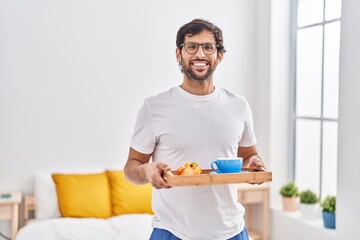 Handsome latin man eating breakfast on the bed smiling with a happy and cool smile on face. showing teeth.