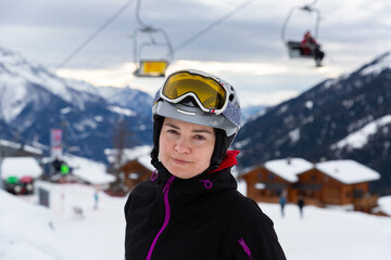 Fototapeta na wymiar Young woman spending winter holidays in ski resort, posing in ski helmet and goggles outdoors with snow covered mountains in background