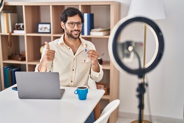 Handsome latin man recording bitcoin tutorial with smartphone at home smiling happy and positive,...