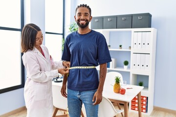 Obraz premium Dietitian at the clinic using tape measure measuring waist of client looking positive and happy standing and smiling with a confident smile showing teeth