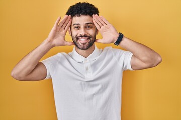 Arab man standing over yellow background smiling cheerful playing peek a boo with hands showing face. surprised and exited