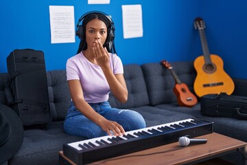 African american woman with braids playing piano keyboard at music studio covering mouth with hand,...