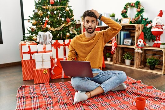 Arab young man using laptop sitting by christmas tree crazy and scared with hands on head, afraid and surprised of shock with open mouth