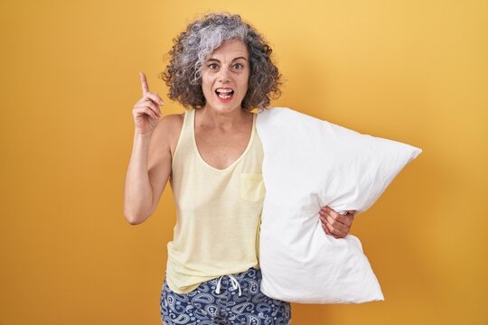 Middle age woman with grey hair wearing pijama hugging pillow pointing finger up with successful idea. exited and happy. number one.