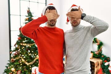 Young gay couple standing by christmas tree wearing hat covering eyes with hand, looking serious and sad. sightless, hiding and rejection concept