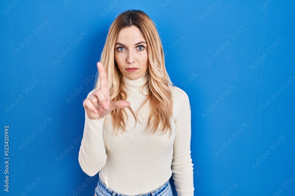 Sticker young caucasian woman standing over blue background pointing with finger up and angry expression, sh