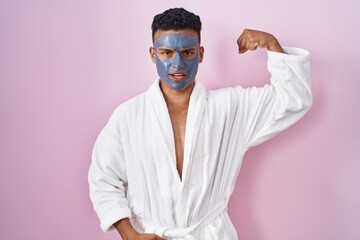 Young hispanic man wearing beauty face mask and bath robe strong person showing arm muscle,...