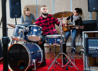 Expressive cheerful smiling drummer with his bandmates practicing in rehearsal room