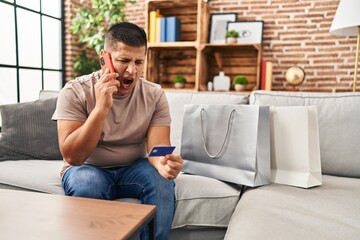 Fototapeta premium Hispanic young man doing payment with credit card on the phone angry and mad screaming frustrated and furious, shouting with anger. rage and aggressive concept.