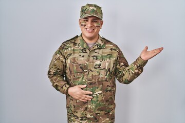 Hispanic young man wearing camouflage army uniform smiling cheerful presenting and pointing with palm of hand looking at the camera.