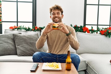 Young hispanic man eating hamburger sitting by christmas decor at home