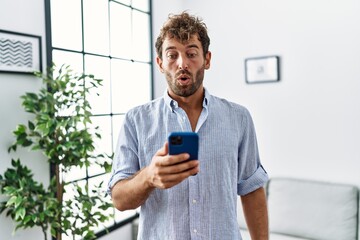 Young handsome man using smartphone at home scared and amazed with open mouth for surprise, disbelief face