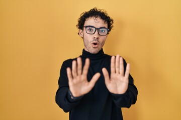 Fototapeta premium Hispanic man standing over yellow background moving away hands palms showing refusal and denial with afraid and disgusting expression. stop and forbidden.