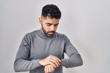 Hispanic man with beard standing over white background checking the time on wrist watch, relaxed and confident