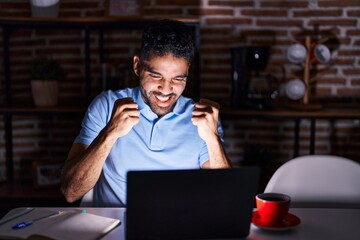 Fototapeta na wymiar Hispanic man with beard using laptop at night very happy and excited doing winner gesture with arms raised, smiling and screaming for success. celebration concept.