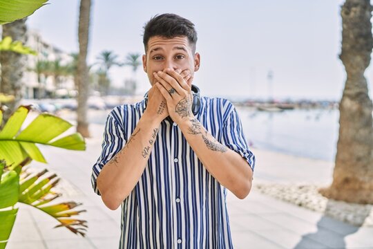 Young handsome man listening to music using headphones outdoors shocked covering mouth with hands for mistake. secret concept.