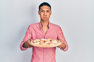 Young african american guy holding tray with cake sweets puffing cheeks with funny face. mouth...