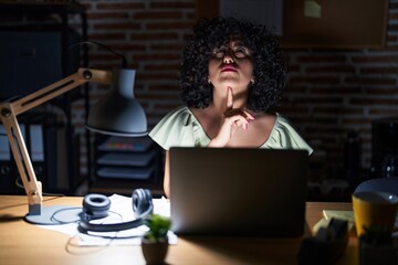 Young brunette woman with curly hair working at the office at night thinking concentrated about...