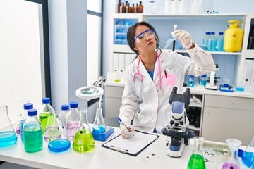 Young chinese woman wearing scientist uniform holding blood test tube writing on document at laboratory