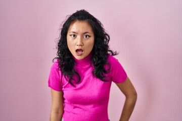 Young asian woman standing over pink background in shock face, looking skeptical and sarcastic, surprised with open mouth