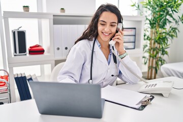 Young hispanic woman wearing doctor uniform talking on the telephone at clinic