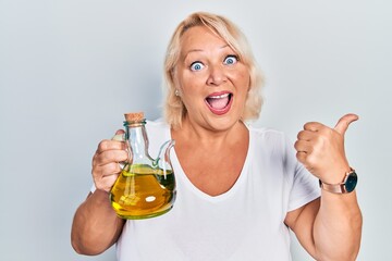 Middle age blonde woman holding olive oil can pointing thumb up to the side smiling happy with open...