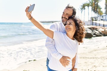 Middle age hispanic couple smiling happy and hugging making selfie by the smartphone at the beach.