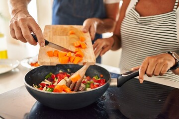 Middle age hispanic couple pouring food on frying pan at kitchen