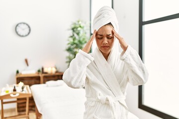 Young brunette woman wearing towel and bathrobe standing at beauty center with hand on head for pain in head because stress. suffering migraine.