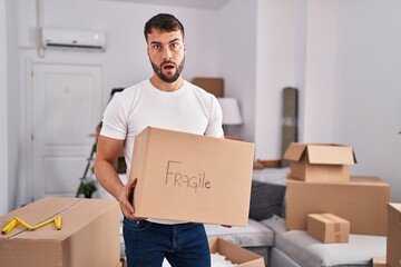 Handsome hispanic man moving to a new home holding fragile box in shock face, looking skeptical and sarcastic, surprised with open mouth