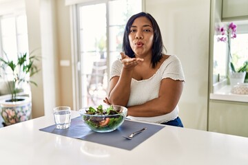 Young hispanic woman eating healthy salad at home looking at the camera blowing a kiss with hand on air being lovely and sexy. love expression.